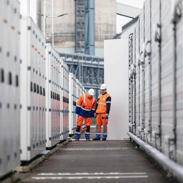 Zwei Arbeiter in orangener Sicherheitskleidung untersuchen Geräte zwischen Reihen von großen weißen Containern auf einer Baustelle.