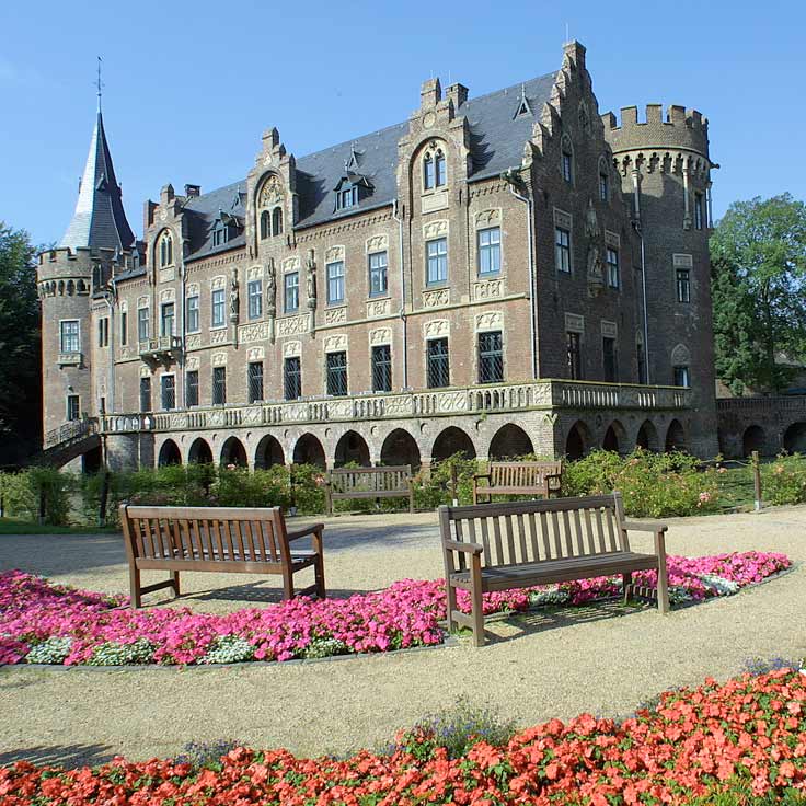 Ein großes historisches Schloss mit turmartiger Architektur, umgeben von bunten Blumenbeeten und Bänken in einem sonnigen Garten.