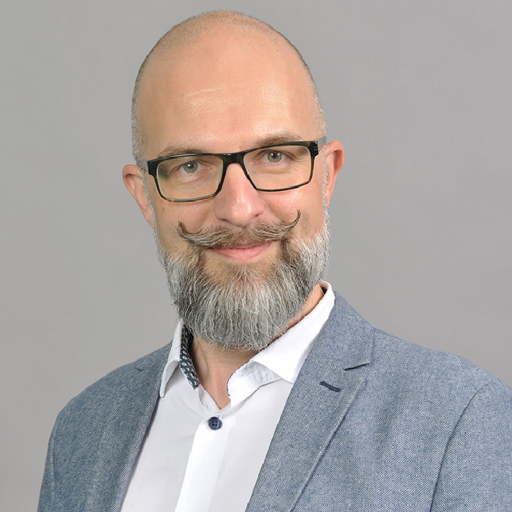 Ein Mann mit grauem Bart trägt ein hellblaues Blazer und ein weißes Hemd. Der Hintergrund ist grau, Portrait von Dr. Jens Wiggershaus.