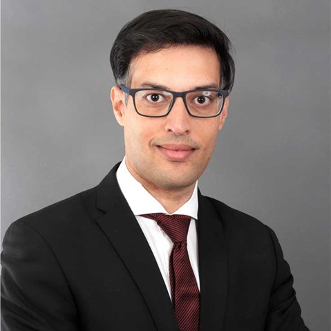A man in a black suit and red tie stands against a grey background, posing in a professional manner. Portrait of Kunal Chandra.