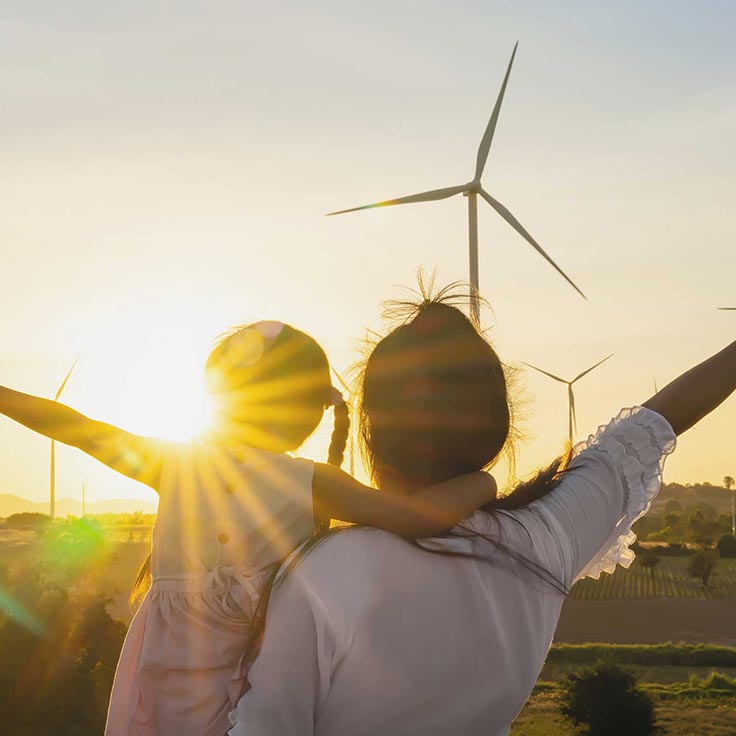 Eine Frau hält ein Kind, während sie bei Sonnenuntergang vor Windkraftanlagen stehen. Die Sonne strahlt hell.
