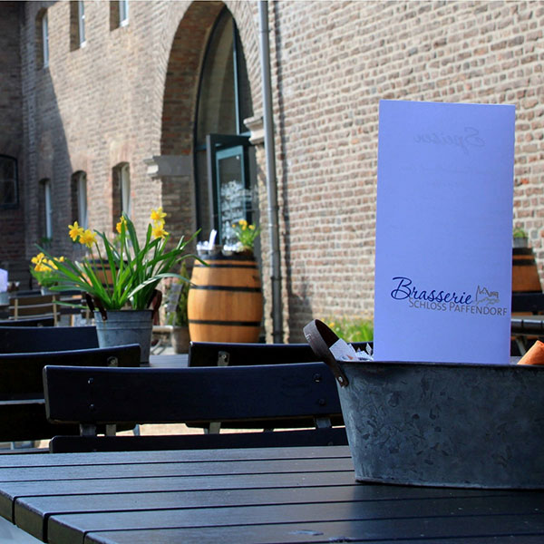 Ein Tisch im Freien mit einer Speisekarte und Blumen in Töpfen vor einer historischen Backsteinmauer.