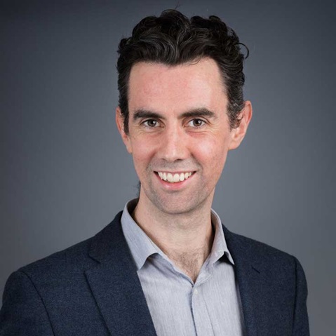 Ein Mann in einem blauen Blazer und einem hellen Hemd steht vor einem grauen Hintergrund, Portrait von Chris Willow.