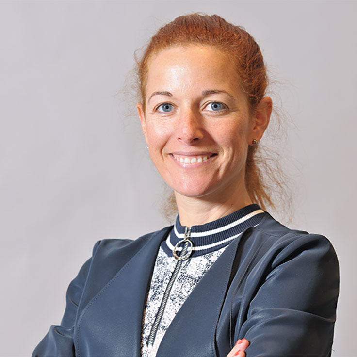 Eine Person mit roten Haaren trägt eine elegante, dunkelblaue Jacke mit einem Reissverschluss und einem gemusterten Oberteil, Portrait von Marta Carroza Diaz.