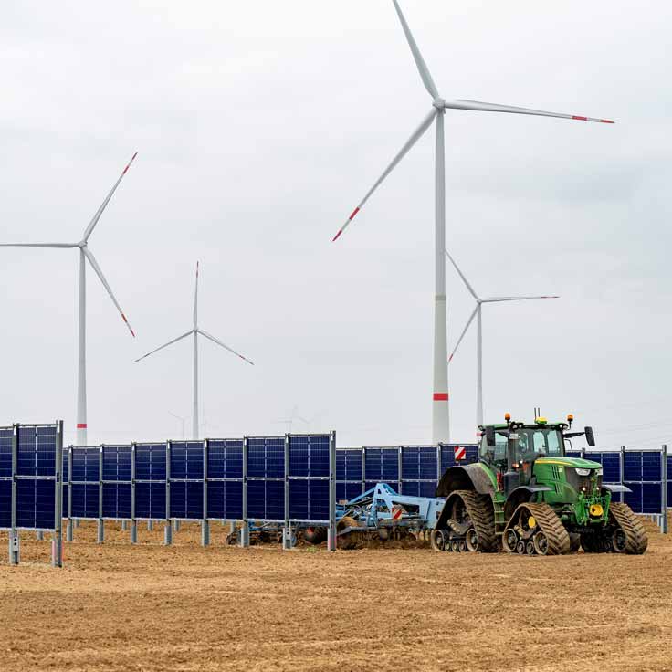 A tractor cultivates soil between solar panels, with wind turbines visible in the background.