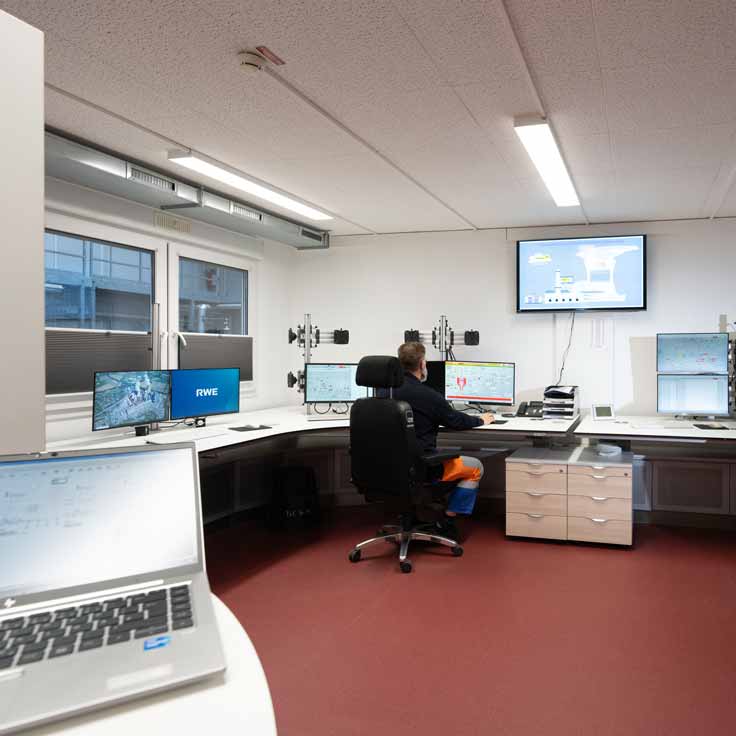 Ein Mann sitzt in einem modernen Kontrollraum, umgeben von mehreren Bildschirmen und Arbeitsstationen.