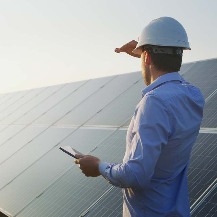 Ein Mann in einem Bauhelm steht vor Solarpanelen und schaut in die Ferne, während er ein Tablet hält.