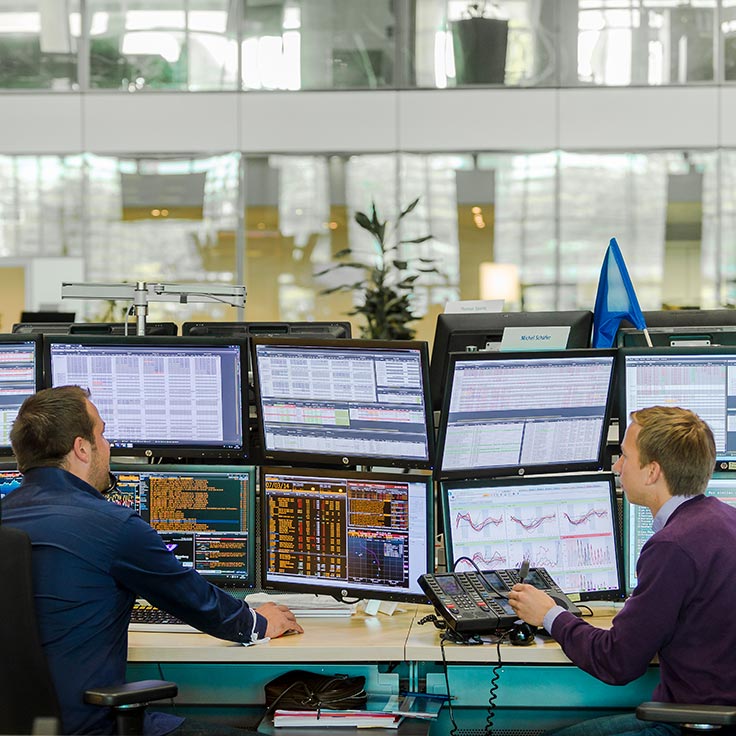 Two traders work at multiple monitors in a modern office, analysing financial data and charts.