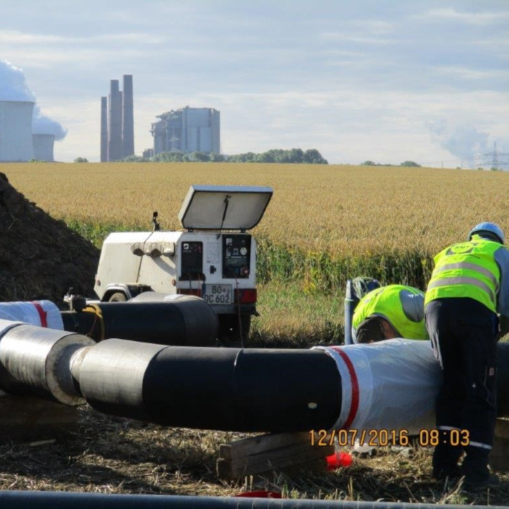 Eine Reihe großer Metallrohre, die parallel zu einem grünen Feld unter einem hellblauen Himmel mit flauschigen Wolken verlaufen.
