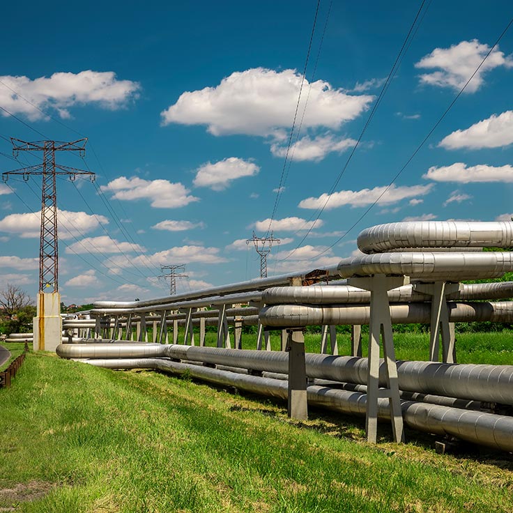 Eine Reihe großer Metallrohre, die parallel zu einem grünen Feld unter einem hellblauen Himmel mit flauschigen Wolken verlaufen.