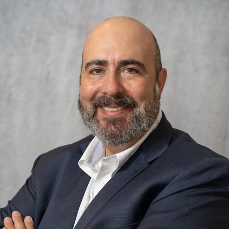 Ein Mann in einem Anzug mit verschränkten Armen vor einem grauen Hintergrund, Kleidung sieht professionell aus. Portrait von Guillermo Schmidhuber.