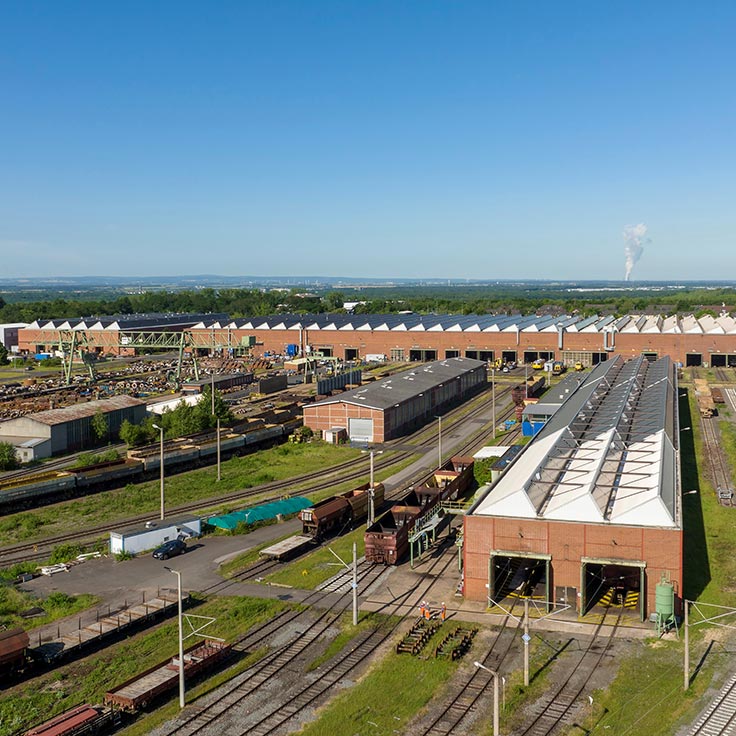 Luftaufnahme eines Industriegebiets mit Lagerhäusern, Bahngleisen und geparkten Waggons unter einem klaren blauen Himmel.