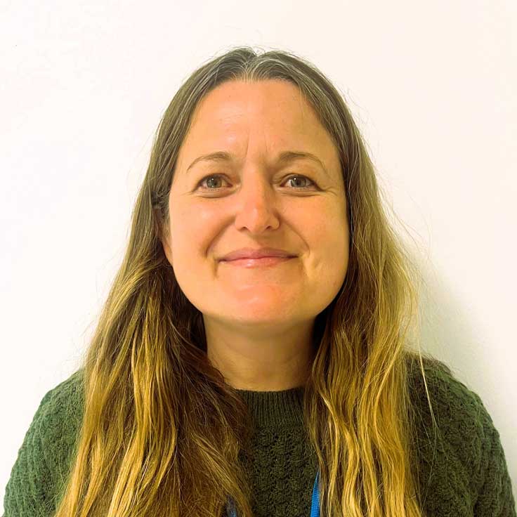 Eine Person mit langen, gewellten Haaren trägt einen grünen Pullover vor einem weißen Hintergrund. Portrait von Joanne Rose.