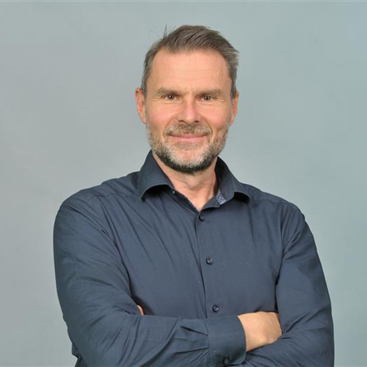 A man in a dark shirt stands with crossed arms in front of a grey background.