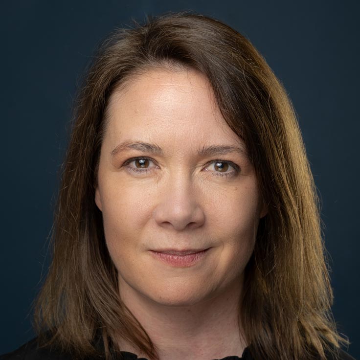 Eine Person mit schulterlangem braunem Haar, die ein schwarzes Oberteil vor einem dunklen Hintergrund trägt. Portrait von Melanie Dreesen.