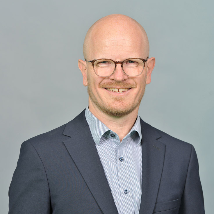 A professional man in a suit and collared shirt poses against a muted grey background. Portrait of Olaf Mönig.