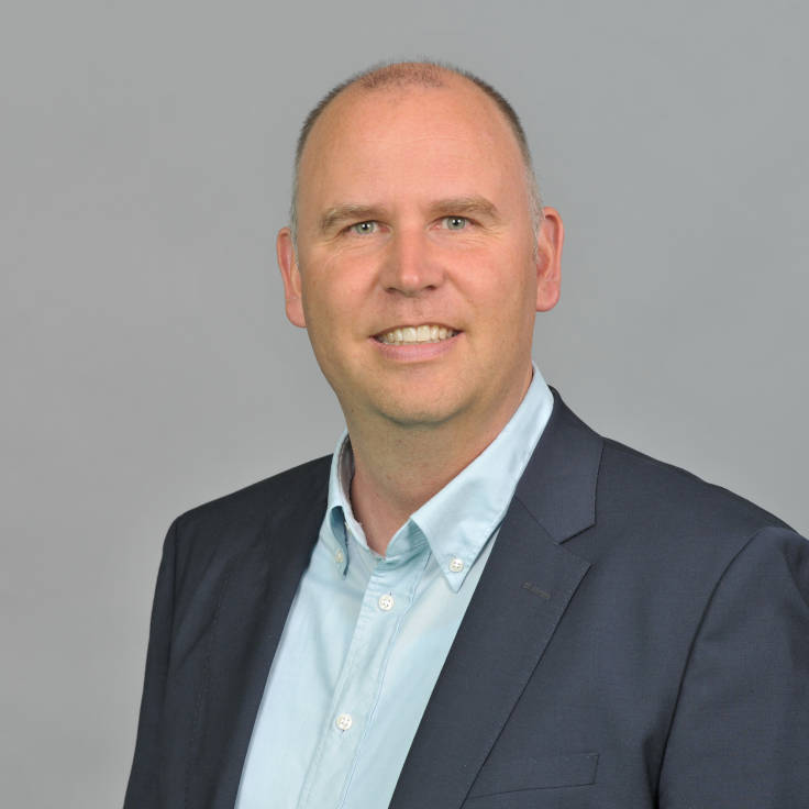 Ein Mann in einem Anzug und einem hellblauen Hemd steht vor einem grauen Hintergrund und vermittelt ein professionelles Erscheinungsbild. Portrait von Philipp Biallass.