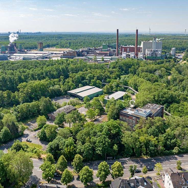 Luftaufnahme eines Industriegebiets, umgeben von Wald und Bäumen, mit mehreren Fabriken im Hintergrund.