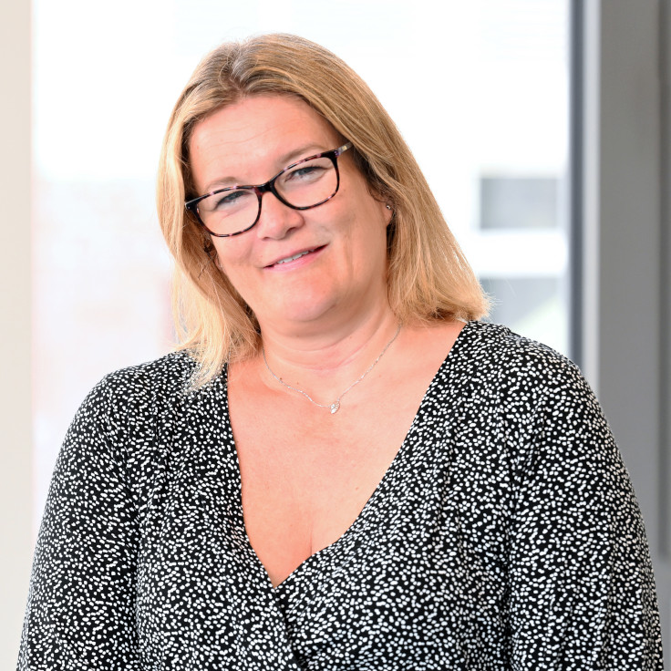 A person with blonde hair is wearing a spotted top and standing in a modern space with large windows. Portrait of Abigail Jerrison.