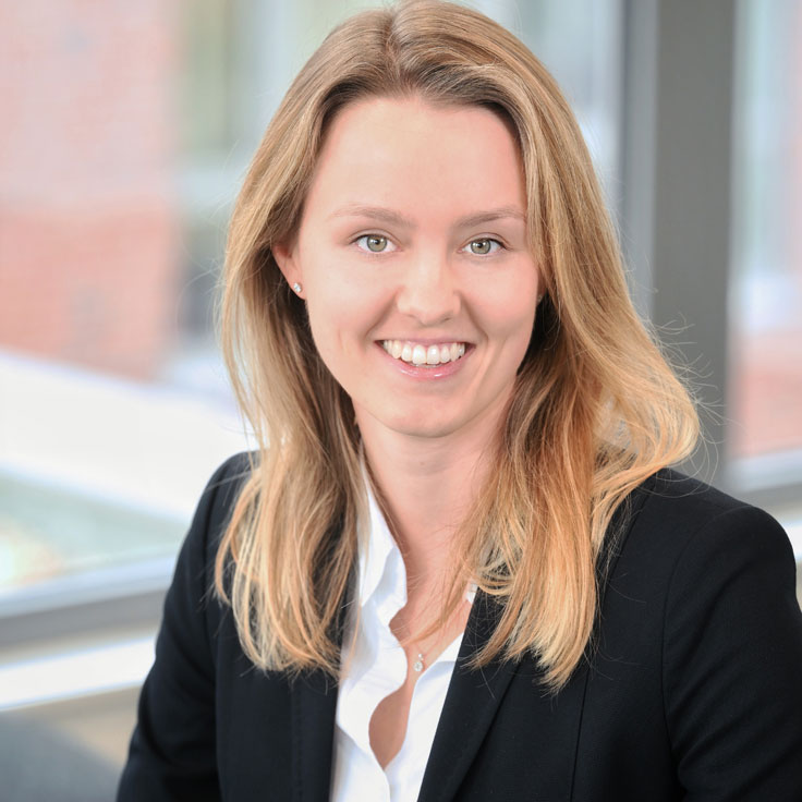 A person with long, blonde hair is wearing a black blazer and a white shirt in a modern office setting. Portrait of Charlotte Mosel.