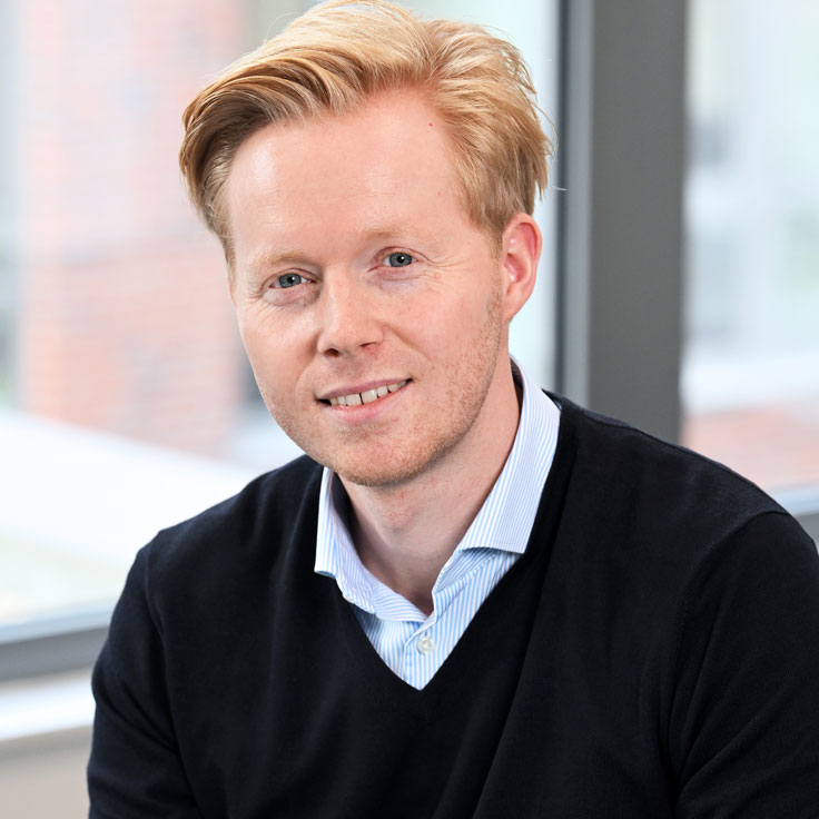 A person with blonde hair is wearing a black sweater and a light blue shirt. There are windows in the background. Portrait of Eric Westphal.