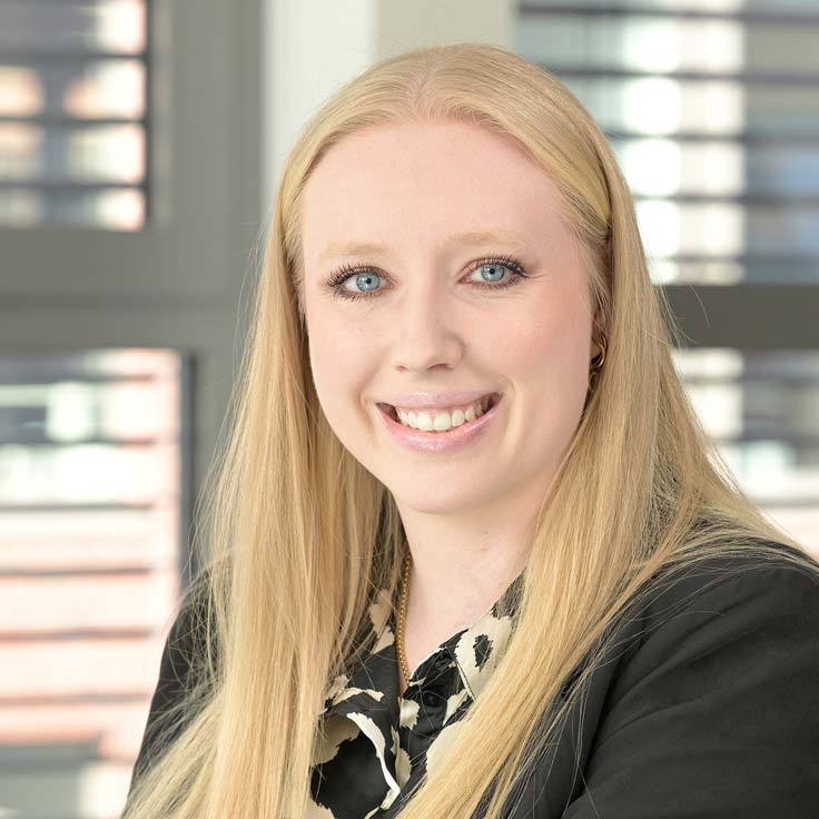A person with long, blonde hair is wearing a black and white blouse and is seated in front of a large window. Portrait of Jacqueline Medelnik.