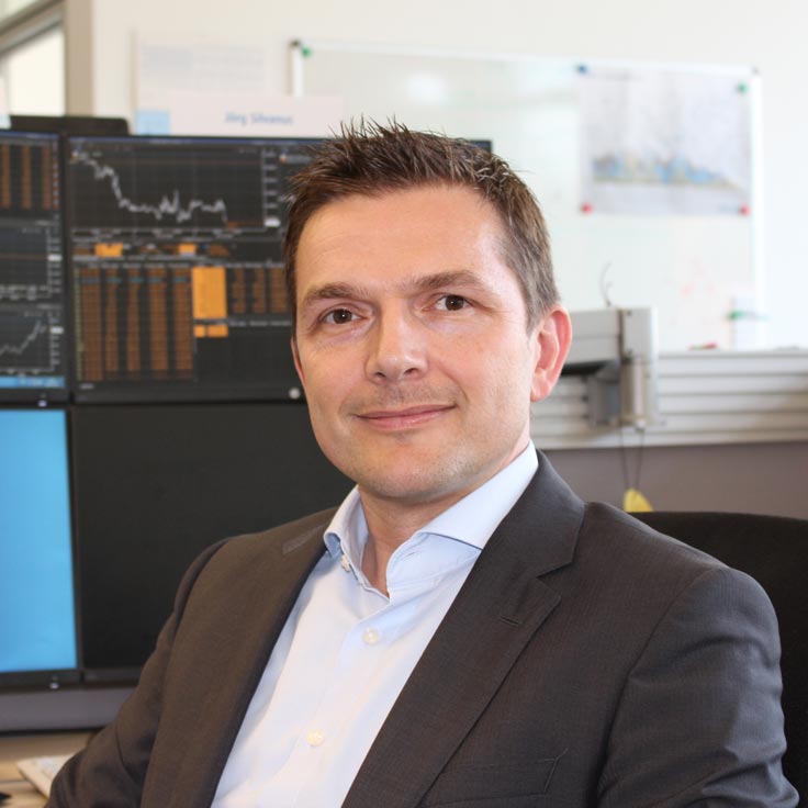 A man in a suit sits in front of multiple computer screens displaying financial data and graphs. Portrait of Jörg Silvanus.