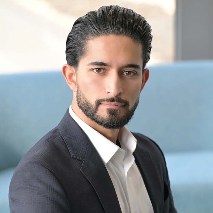 A man in a suit with a white shirt sits on a blue sofa. The background is out of focus. Portrait of Mert Aydin.