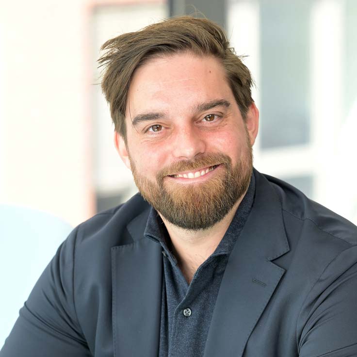 A person in a dark shirt and jacket poses in a modern, bright room. Portrait of Michael Germelmann.