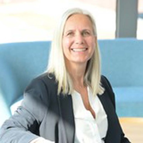 Eine Frau mit langen, blonden Haaren sitzt auf einem blauen Sofa und trägt einen schwarzen Blazer und eine weiße Bluse. Portrait von Nicola Moos.