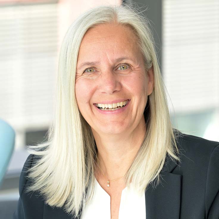 A person with long, blonde hair is wearing a blazer. The background features blurred windows with natural light. Portrait of Nicola Moos.