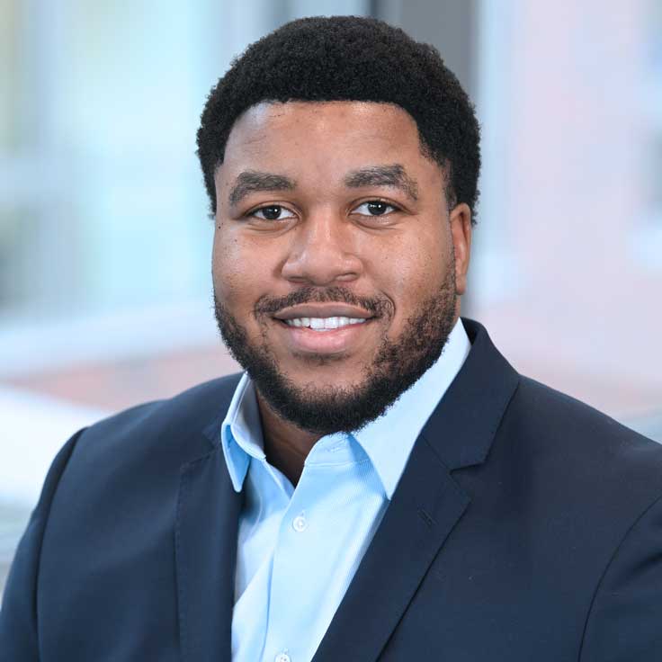 A man is wearing a suit and sitting in a modern setting with a blurred background. Portrait of Praise Sibanda.