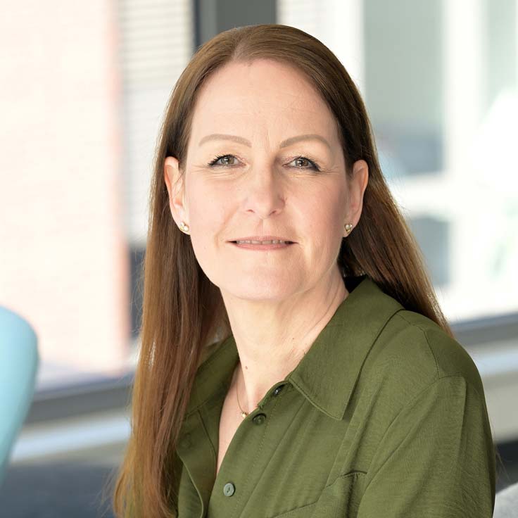 Eine Person mit langen, braunen Haaren trägt ein olivgrünes Hemd und sitzt vor einem großen Fenster. Portrait von Stefanie Nannke.