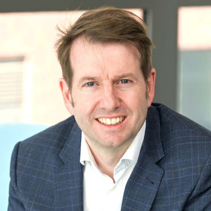A man is wearing a blue suit and a white shirt, sitting at a table with window light in the background. Portrait of Thomas Denny.