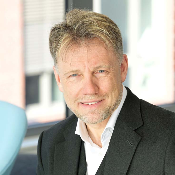 A man in a suit sits in a modern office with a blue chair in the foreground. Portrait of Dr. Burkhard Pahnke.