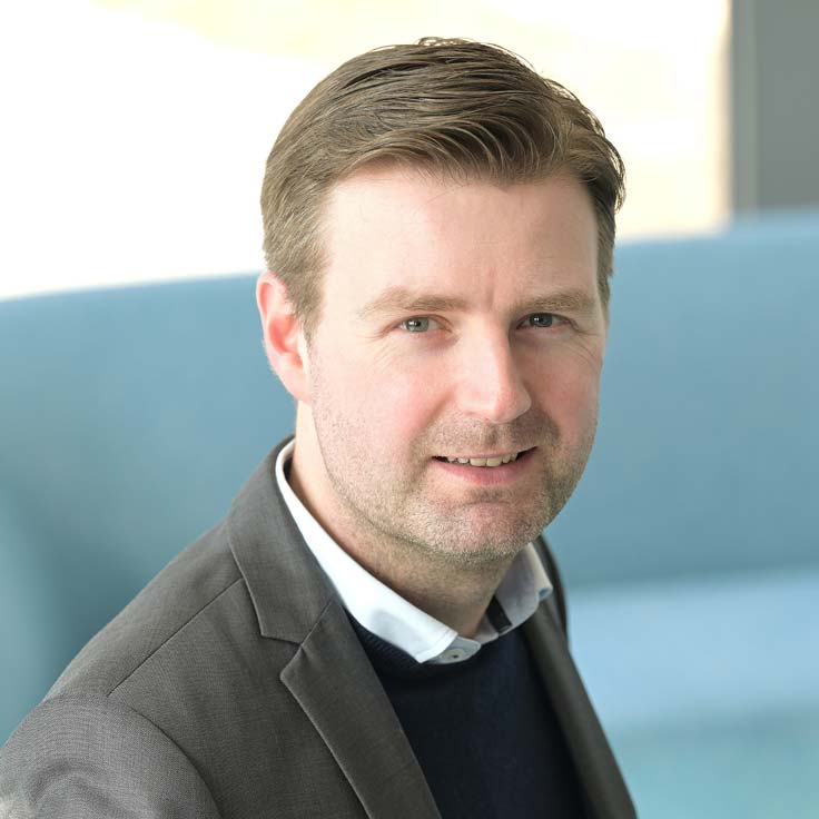 A man in a suit with a blue sweater and a white shirt collar sits in a modern room. Portrait of Jérôme Hördemann.