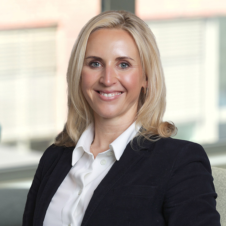 Eine Frau trägt ein schwarzes Sakko und eine weiße Bluse. Der Hintergrund ist unscharf und zeigt Fenster mit Tageslicht. Portrait von Linda Meyerhoff.