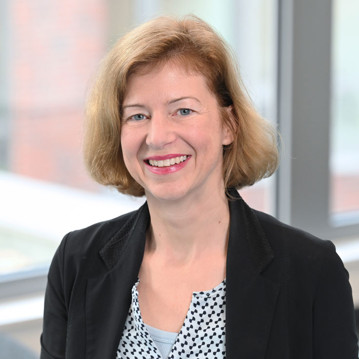 A person with shoulder-length blonde hair wearing a black jacket over a patterned blouse. The background is bright. Portrait of Sabine Rohrbach.