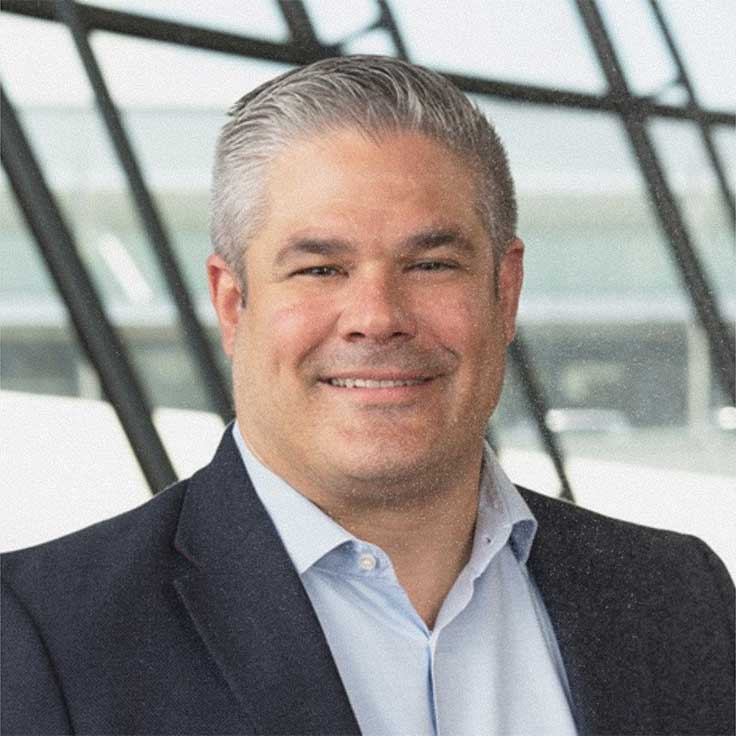 Ein Mann trägt einen Blazer und ein Hemd und ist vor einer modernen Glasarchitektur zu sehen.