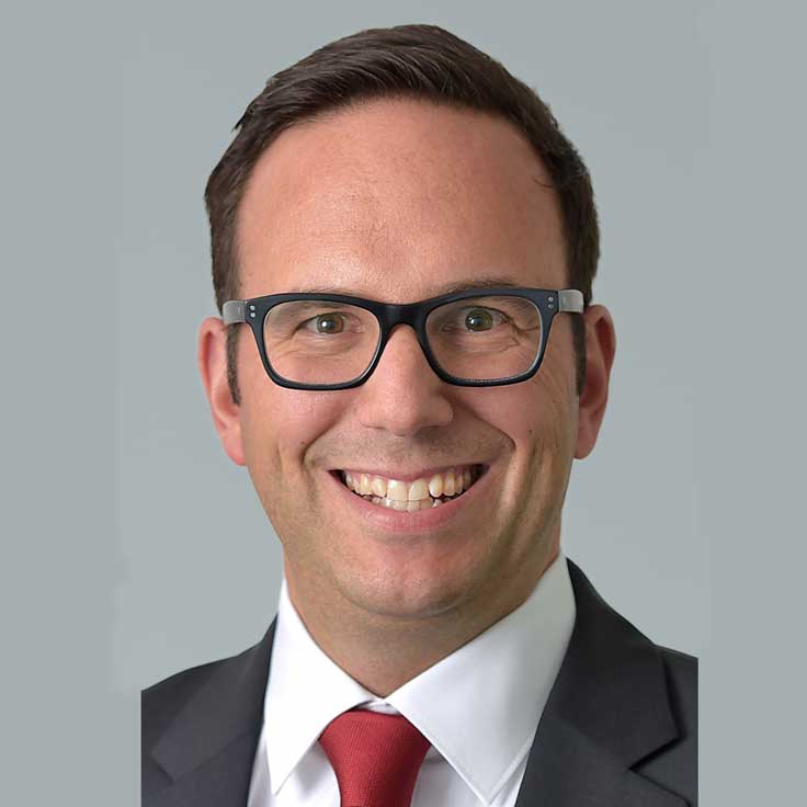 A man in a dark suit with a red tie stands against a grey background, portrait of Michael Veeser.