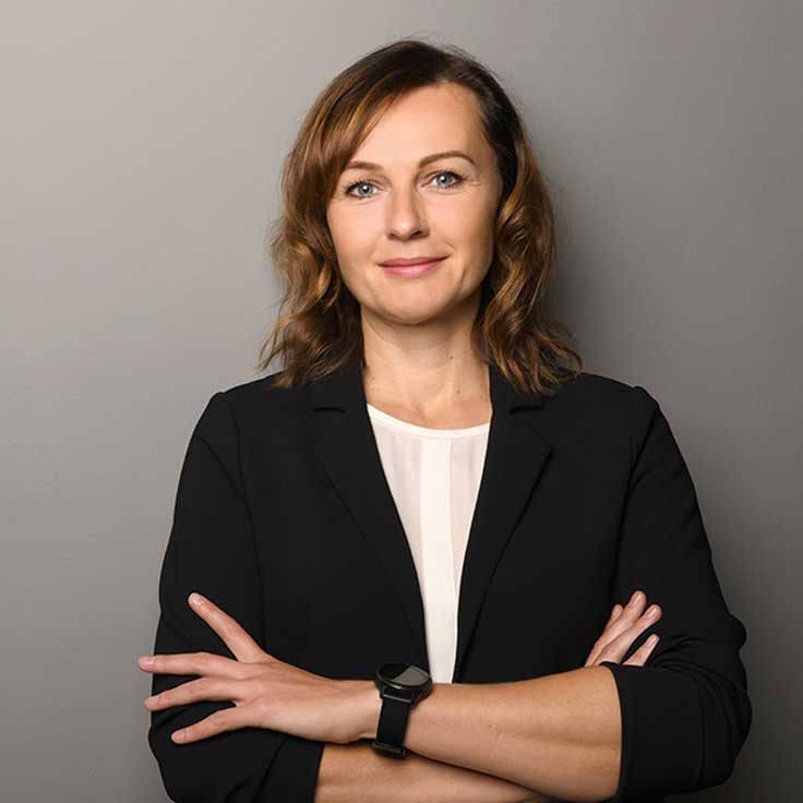 Eine Person mit lockigem, braunem Haar in einem schwarzen Blazer steht mit verschränkten Armen vor einem grauen Hintergrund, Portrait von Monika Witka.