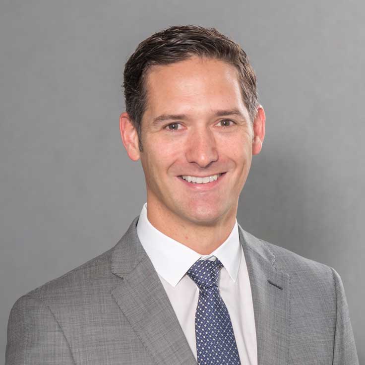 A man in a grey suit with a tie and a white shirt, set against a grey background, portrait of Steven Pryor.
