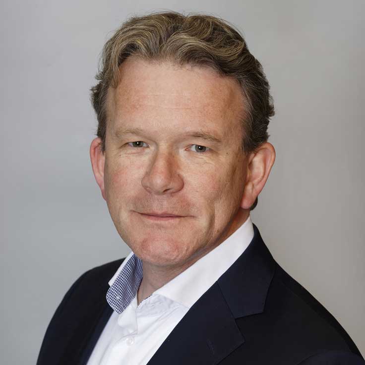 A man in a suit with blonde hair, wearing a white shirt and a blue blazer, against a grey background.