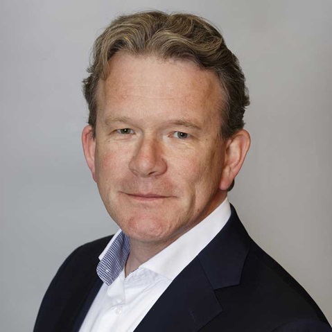 A man in a suit with blonde hair, wearing a white shirt and a blue blazer, against a grey background.