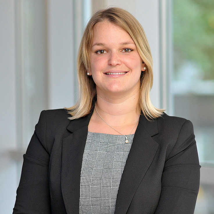 Eine Person in einem Schwarzanzug mit blonden Haaren, die vor einem hellen Hintergrund steht. Portrait von Anna-Lena Meyer.