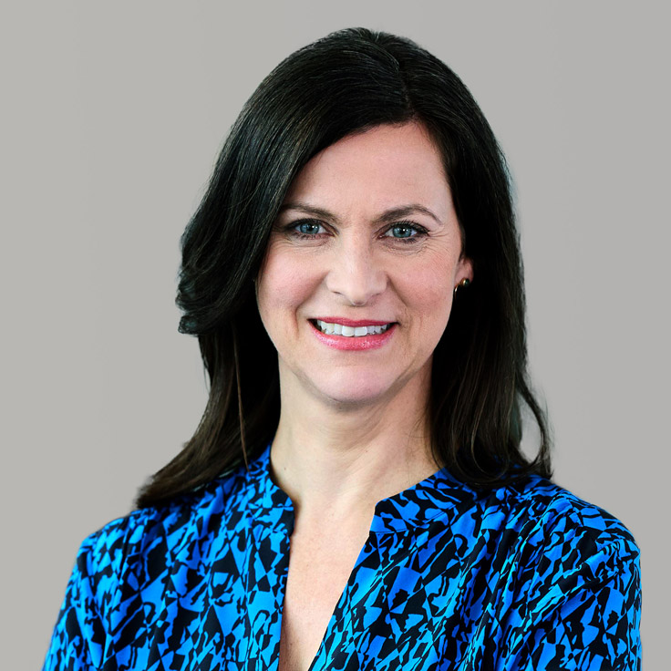 Eine Person mit langen dunklen Haaren trägt eine blau gemusterte Bluse. Der Hintergrund ist grau. Portrait von Birgit Hiller.