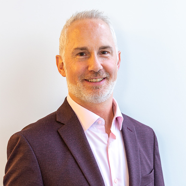 A smiling man with short gray hair and a trimmed beard wears a dark burgundy blazer and a light pink shirt against a pale wall. Portrait of Chris Bauer.