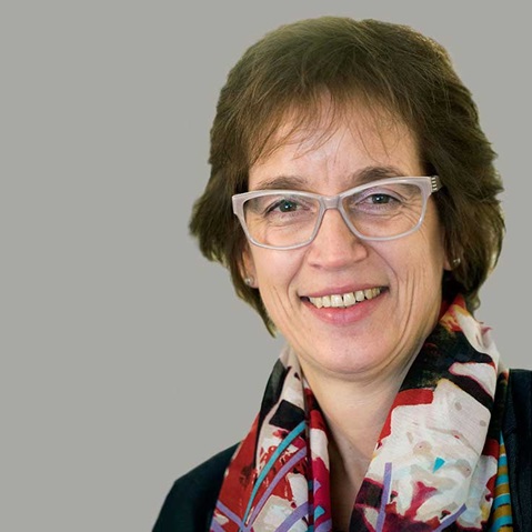 A person with hair wearing a colourful scarf in front of a grey background. Portrait of Dagmar Butz.