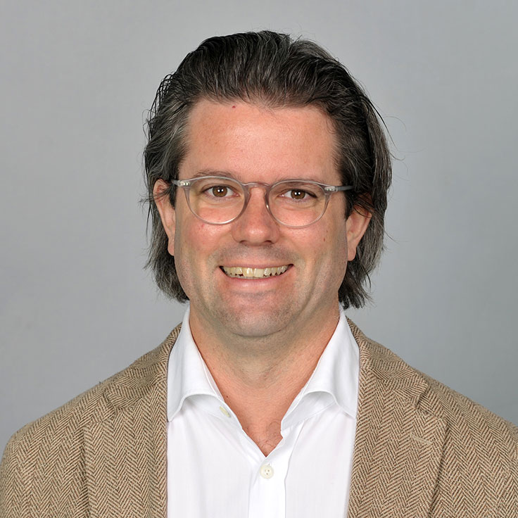 A man is wearing a brown blazer over a white shirt. The background is neutral and grey. Portrait of Heiko Willenberg.
