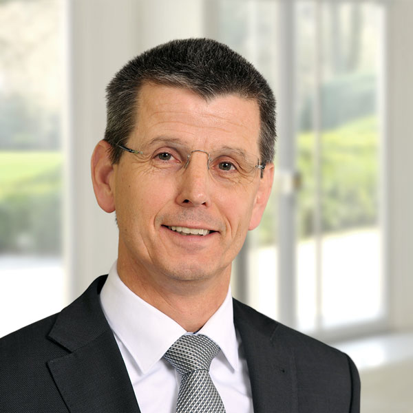 Ein Mann in einem Anzug mit einem grauen Hemd und einer gestreiften Krawatte steht vor einem modernen Fenster. Weiche, grüne Landschaft im Hintergrund. Portrait von lars Kulik.